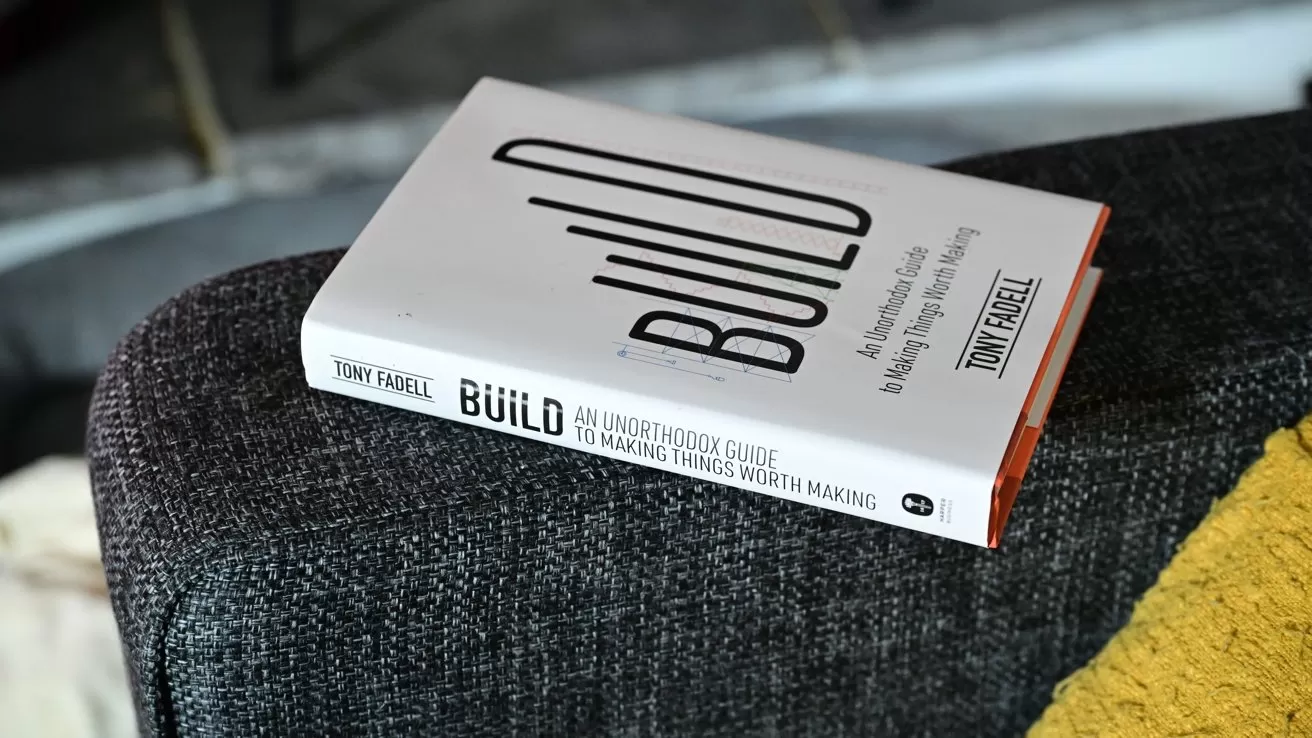 Hardcover book titled Build by Tony Fadell rests on a dark fabric couch near a yellow cushion, viewed at an angle showing the front cover and spine