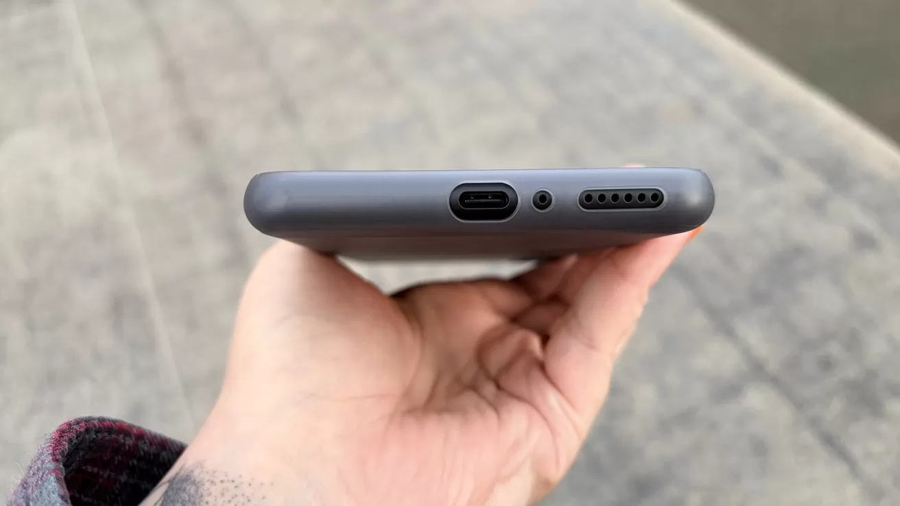 Person holding a smartphone with visible USB-C charging port, headphone jack, and speaker grille on a gray surface background.