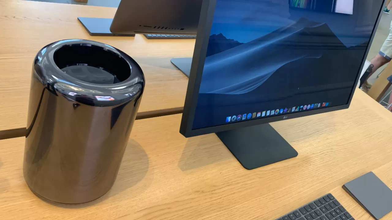 Sleek Apple desktop setup on wooden table with cylindrical black Mac Pro tower next to a large monitor, keyboard, and trackpad in a modern retail store environment