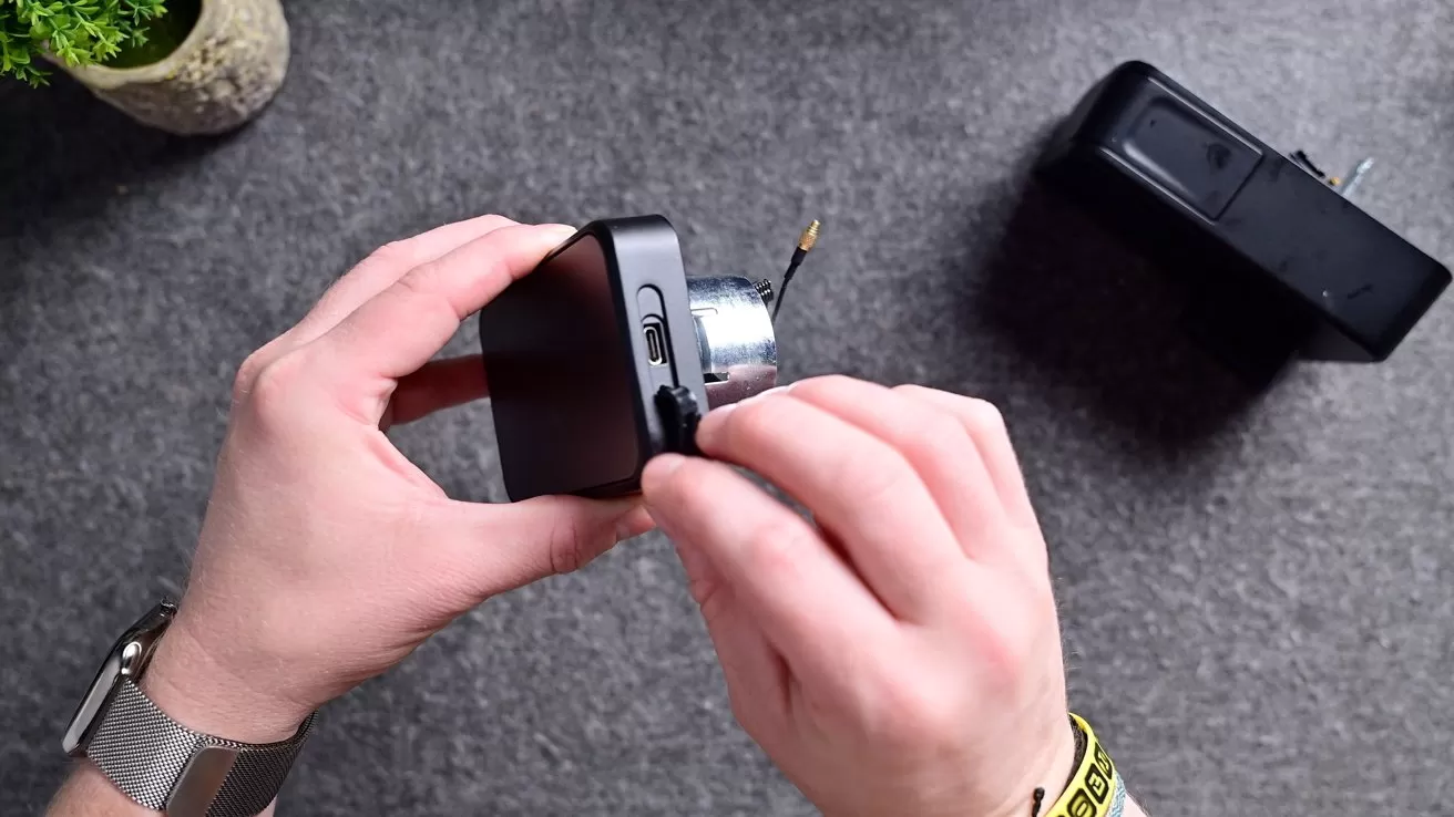 Hands assembling a small black electronic device on a gray table, inserting a cable or connector, with another larger black gadget nearby and a small potted plant in the corner