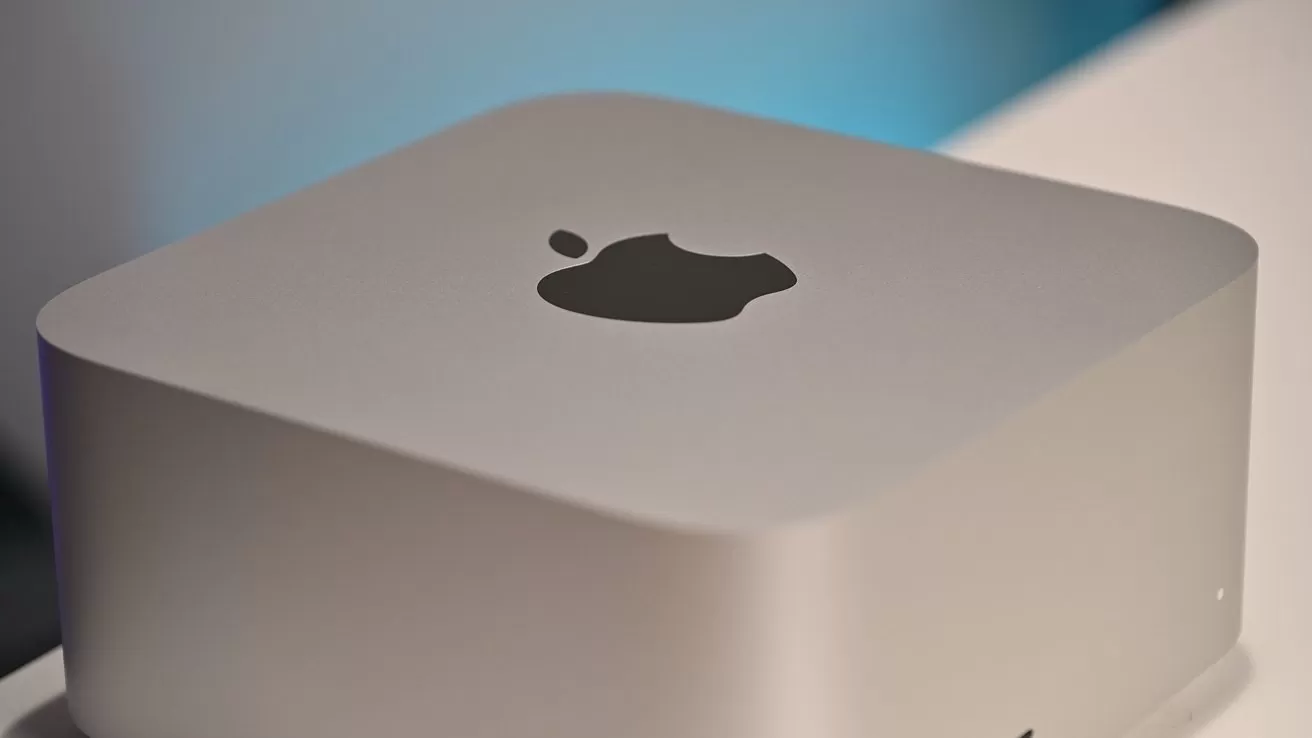 Silver Apple Mac mini desktop computer on a white surface, showing the top with the black Apple logo and softly lit blurred blue background