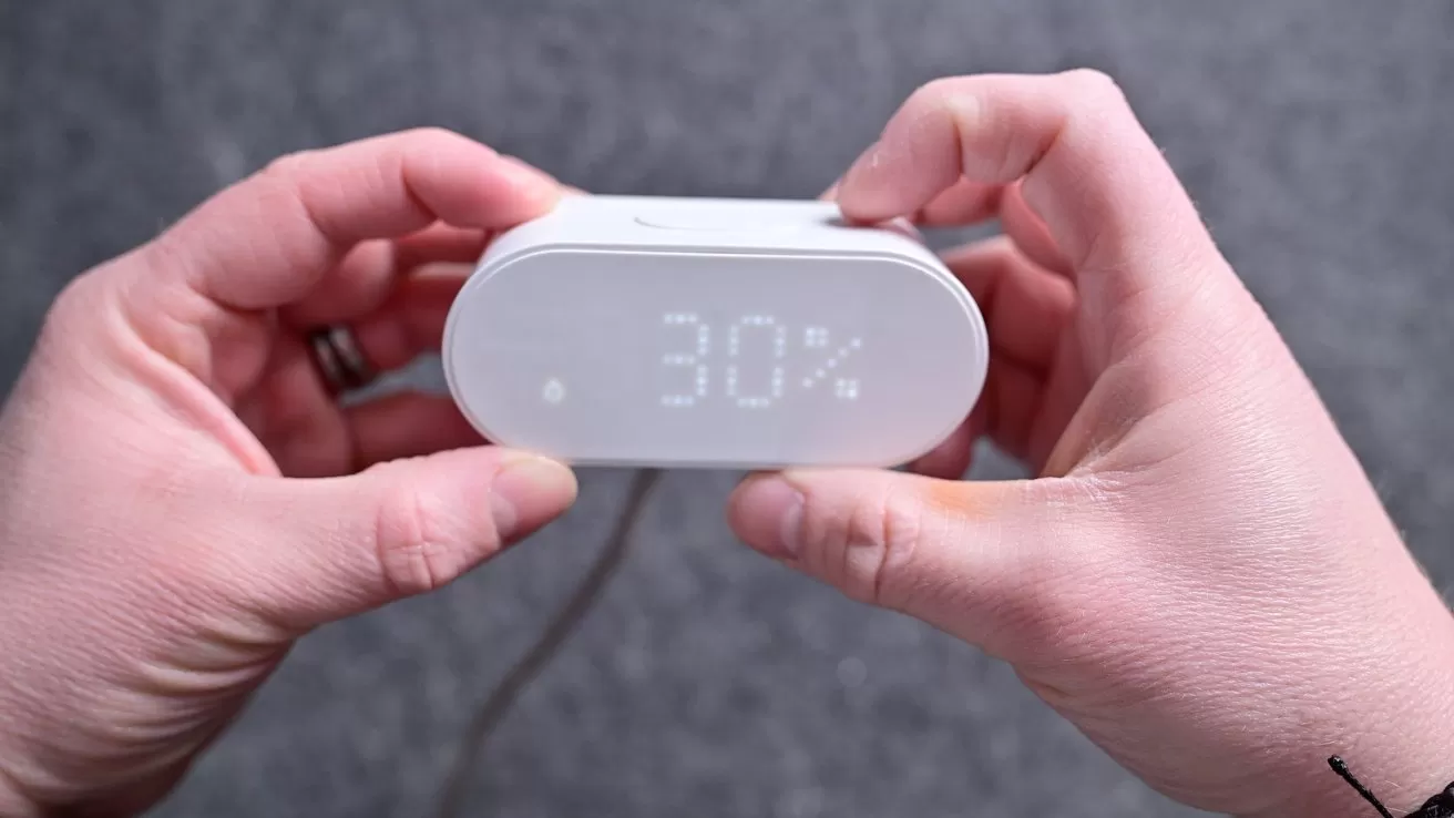 Two hands hold a small white digital device displaying 30 percent on a soft gray surface, with a cable extending from the back