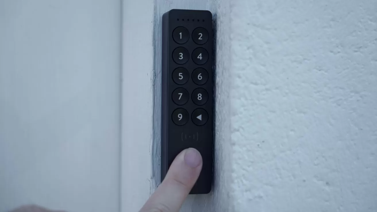 Finger pressing a button on a black numeric keypad mounted on a white wall, suggesting access control or security entry system near a doorway