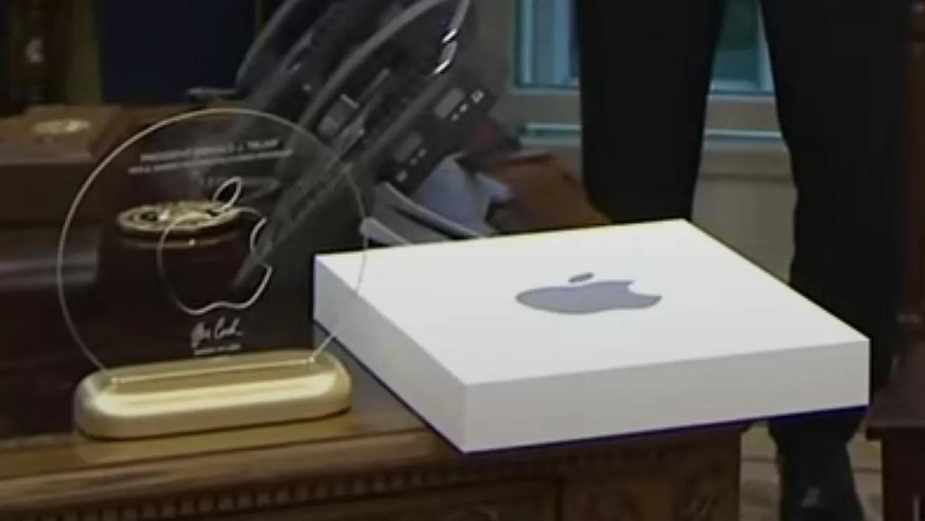 Clear acrylic Apple-shaped award on a gold base beside a white Apple-branded box, resting on an ornate wooden desk with blurred office background