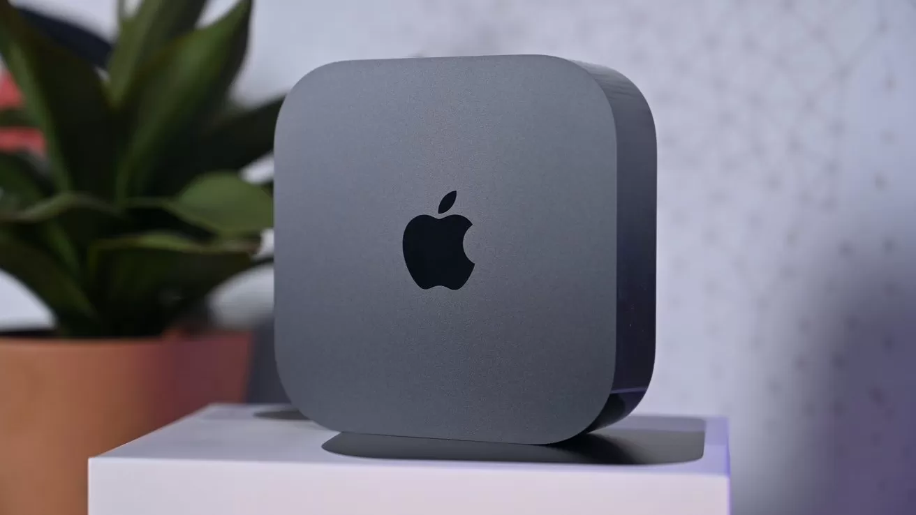 Space gray Apple Mac Mini computer on a white surface, showing the black Apple logo, with a blurred potted plant and patterned wall in the background