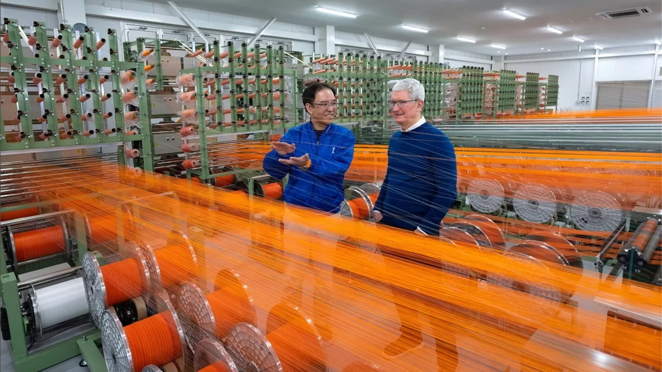 Two men in blue sweaters stand talking inside a brightly lit textile factory, surrounded by fast-moving orange threads and large spools on complex weaving machinery
