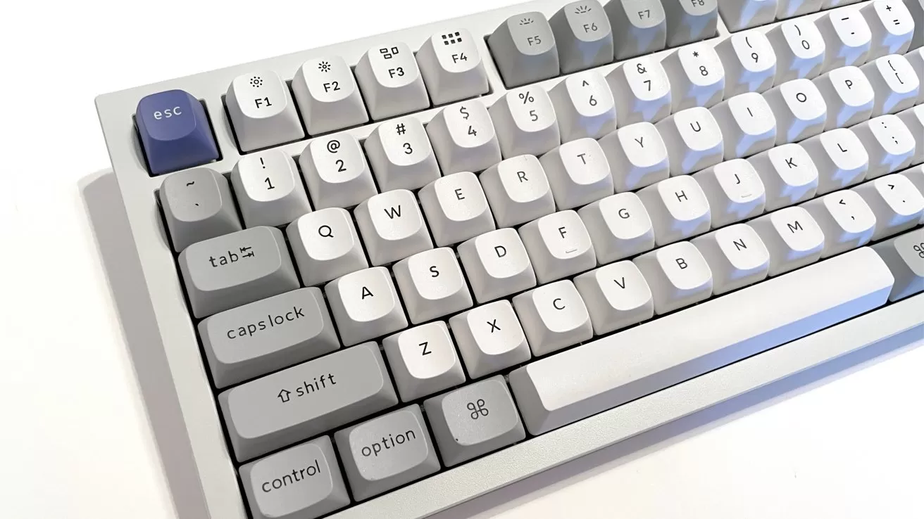 Compact mechanical keyboard with light gray and white keys, dark gray modifier keys, and a single dark blue Escape key in the top left corner on a white surface