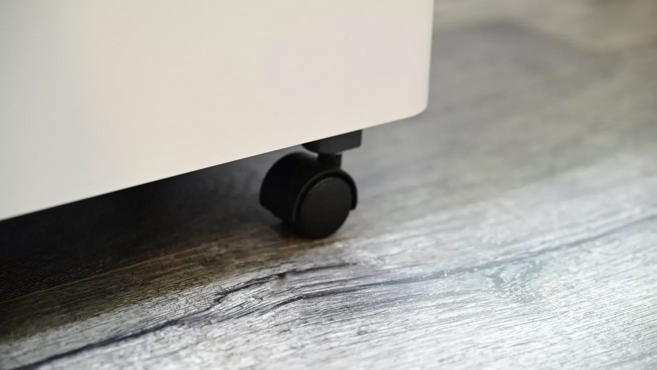 Small black caster wheel under a white piece of furniture resting on light gray wood flooring, viewed close up near the floor level