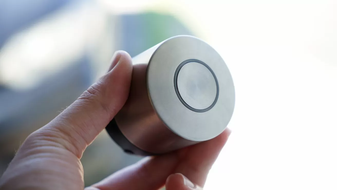 Hand holding a small cylindrical metallic smart lock or knob with a circular button on the end, shown close up against a softly blurred, bright background