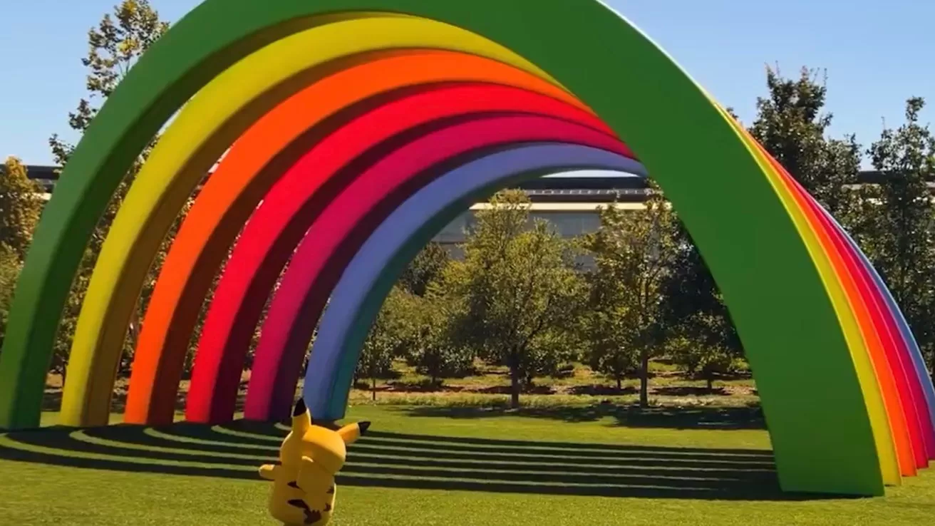 Large colorful rainbow arch sculpture in a sunny park with green grass, trees in the background, and a small Pikachu character standing playfully in the foreground