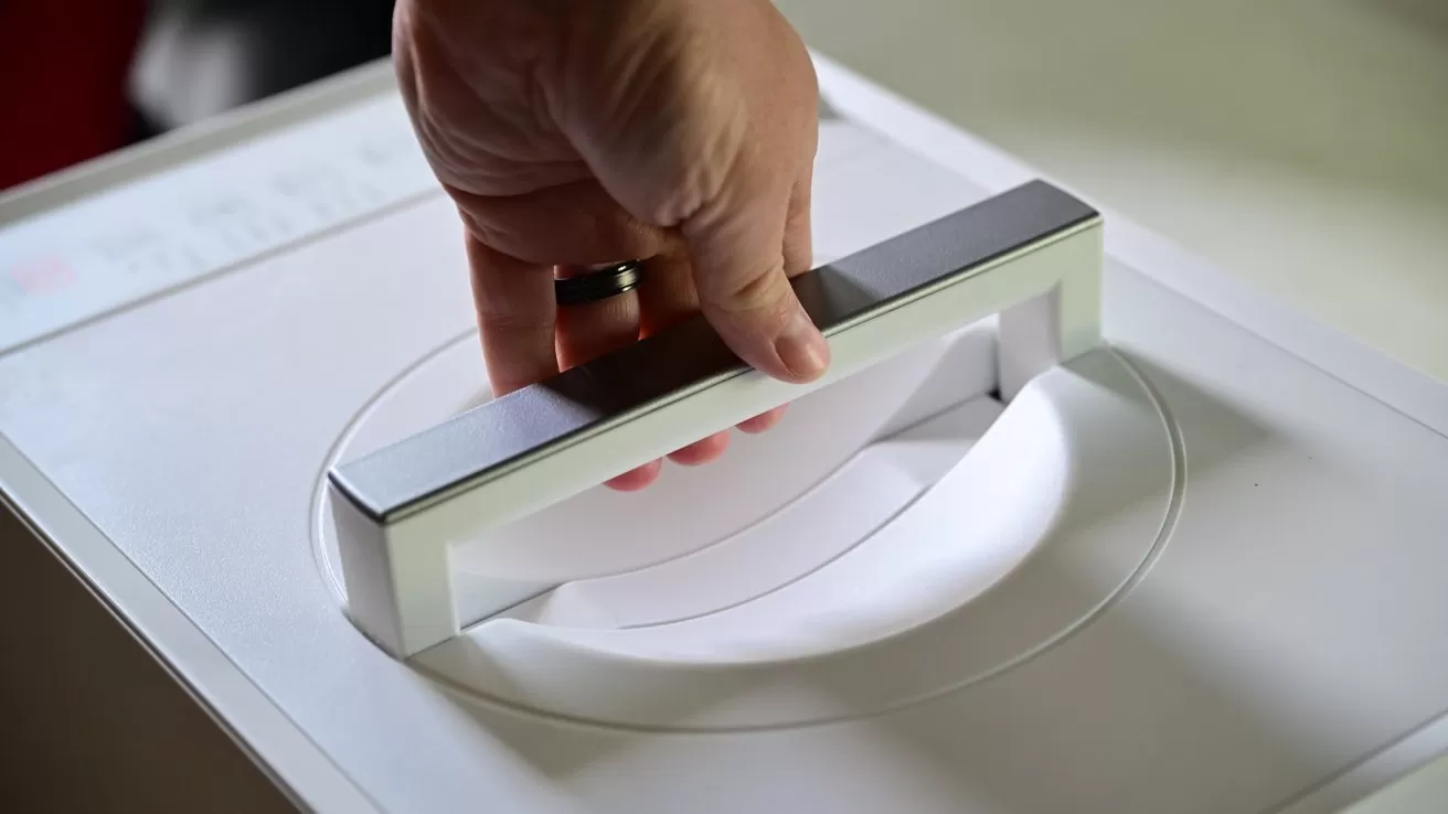 Hand gripping a rectangular handle on top of a white household appliance, possibly a washing machine or dryer, preparing to open the lid
