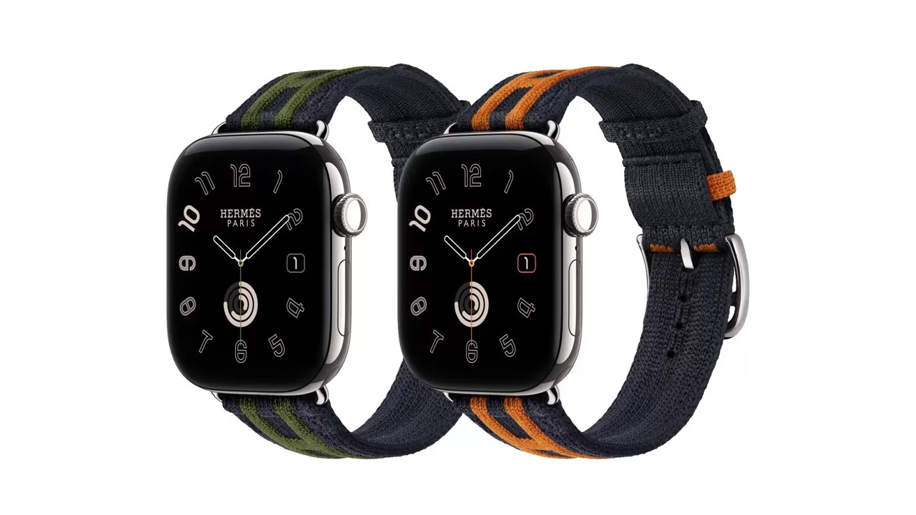Two Apple Watch Hermes models with black faces and dark fabric bands, one featuring green stripes and the other orange stripes, shown side by side on a white background
