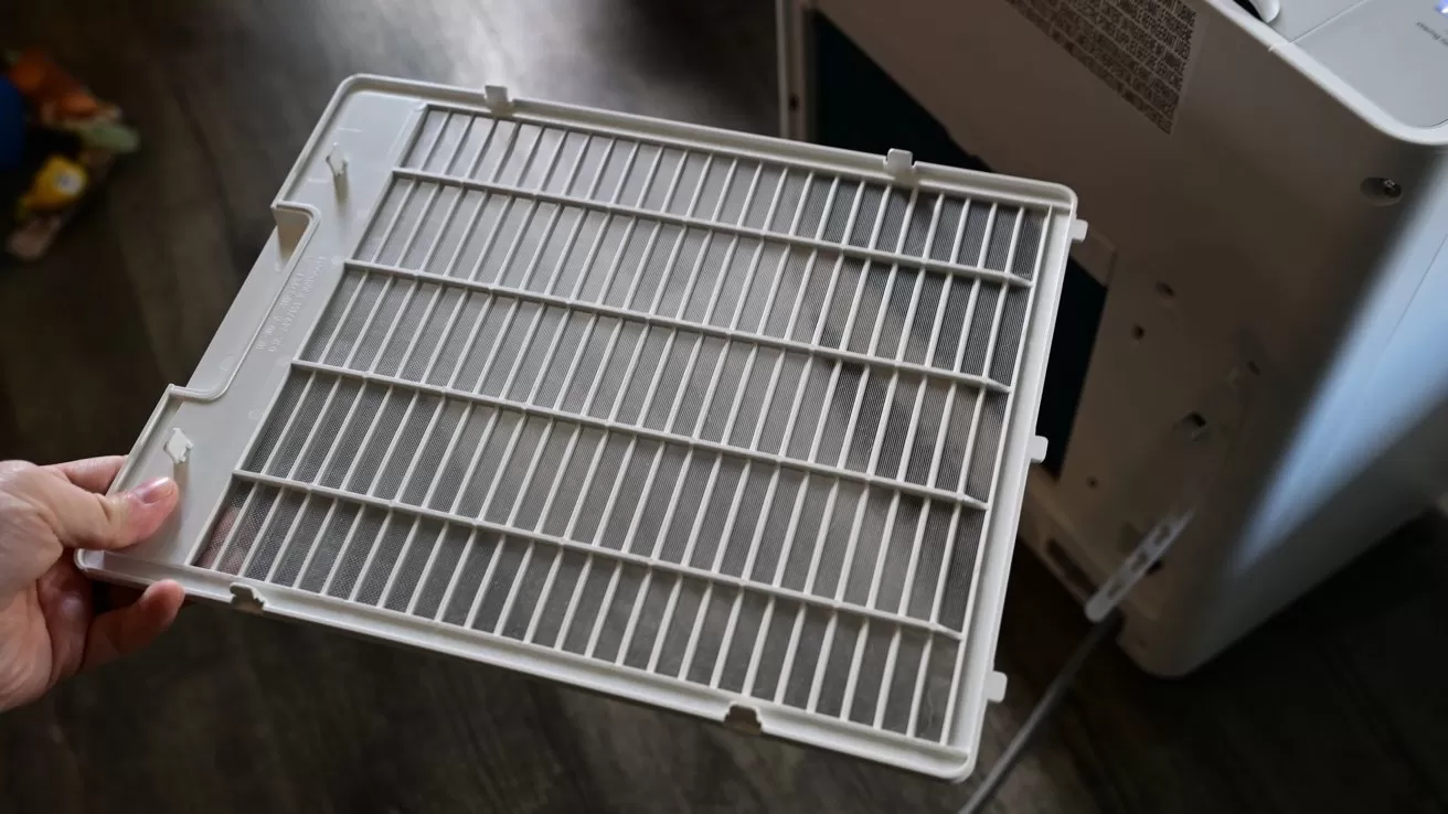 Hand holding a rectangular plastic air filter grate removed from a white household appliance, likely an air purifier or dehumidifier, over a dark wood floor