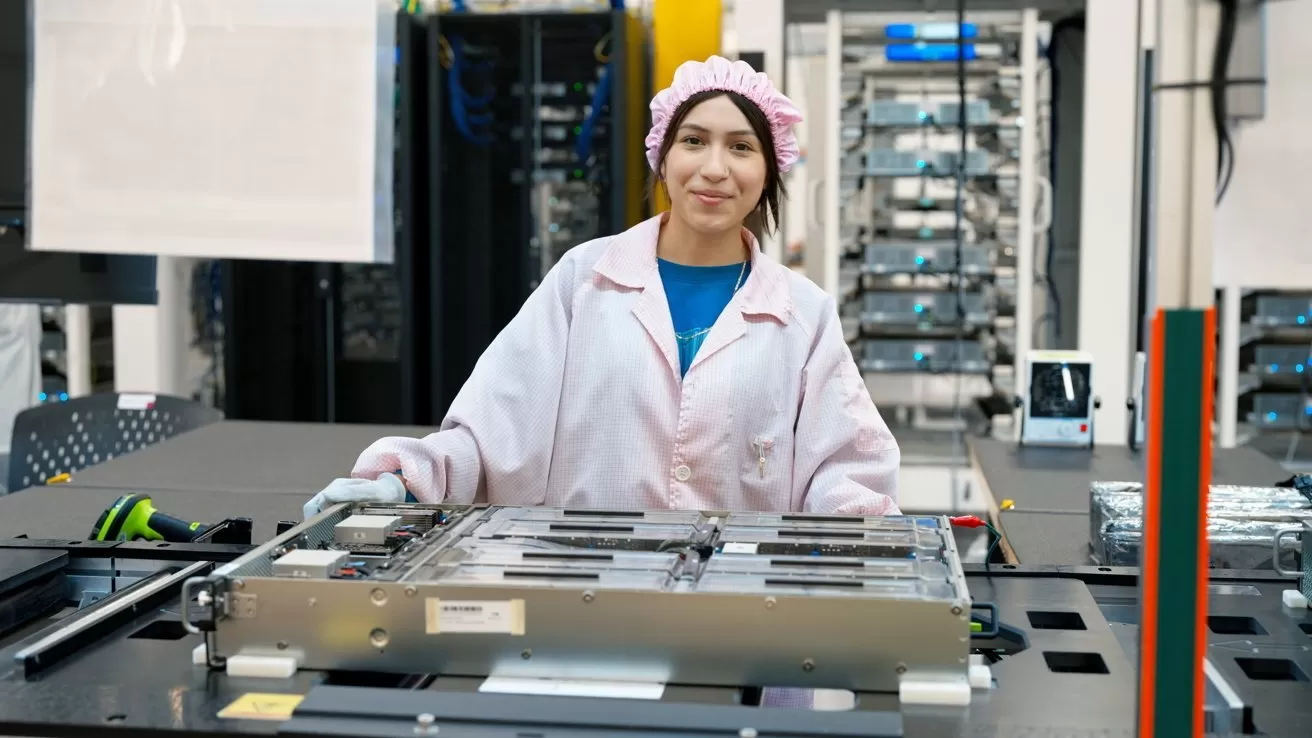 Female technician in a pink lab coat and cap working at an electronics assembly station, handling a large circuit board in a clean, modern factory with server racks in the background