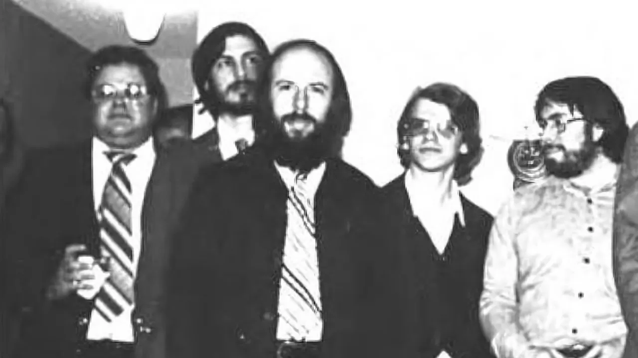 Black and white photo of five men standing close together indoors, most wearing suits and ties, with serious or neutral expressions, facing the camera in a casual group pose