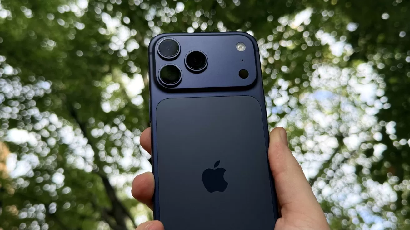 Hand holding a dark blue iPhone outdoors, showing the back with dual camera lenses and Apple logo, against a blurry background of green tree leaves and bright sky.