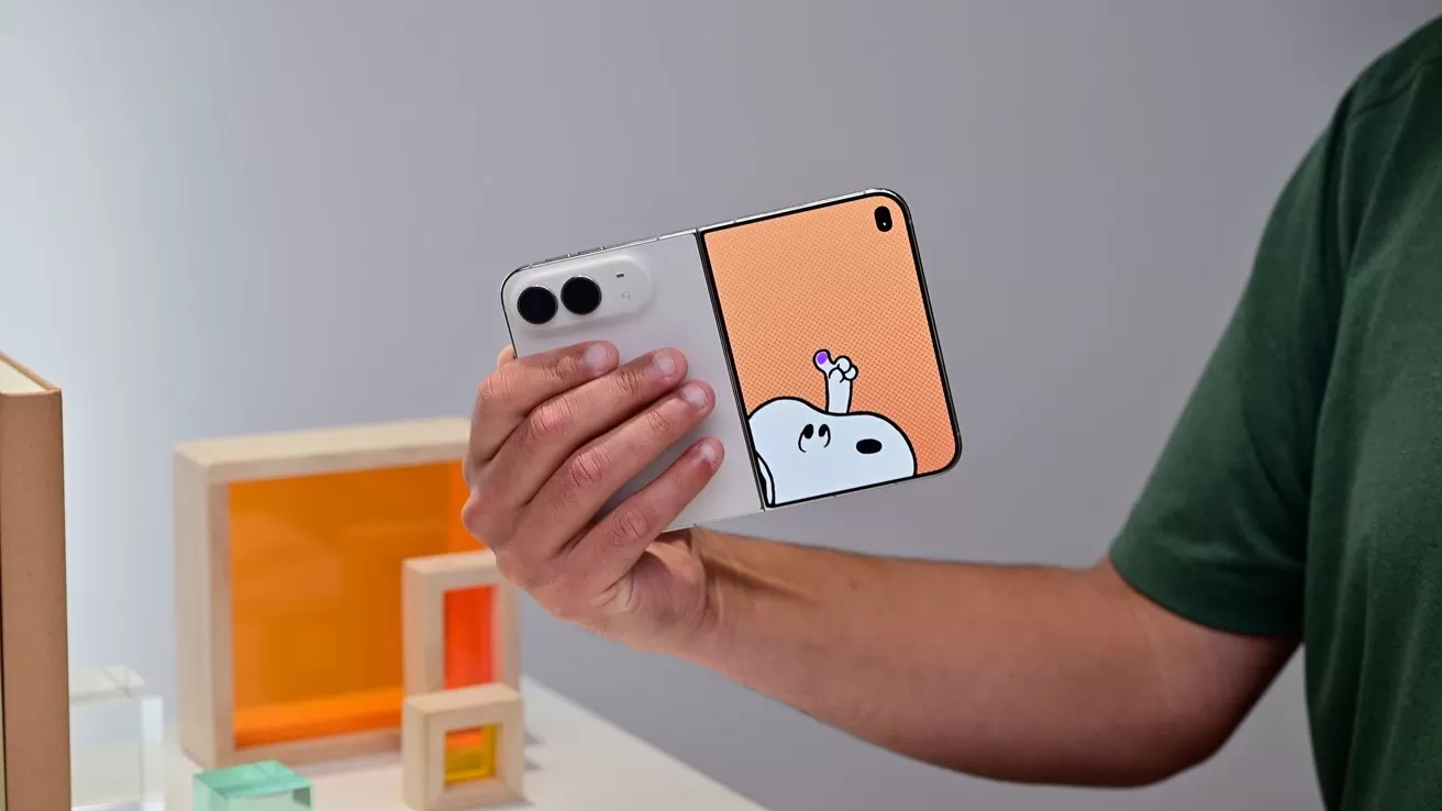 Person holding a foldable smartphone with a playful cartoon character on the unfolded screen, standing indoors near colorful geometric decor blocks and a plain light-colored wall