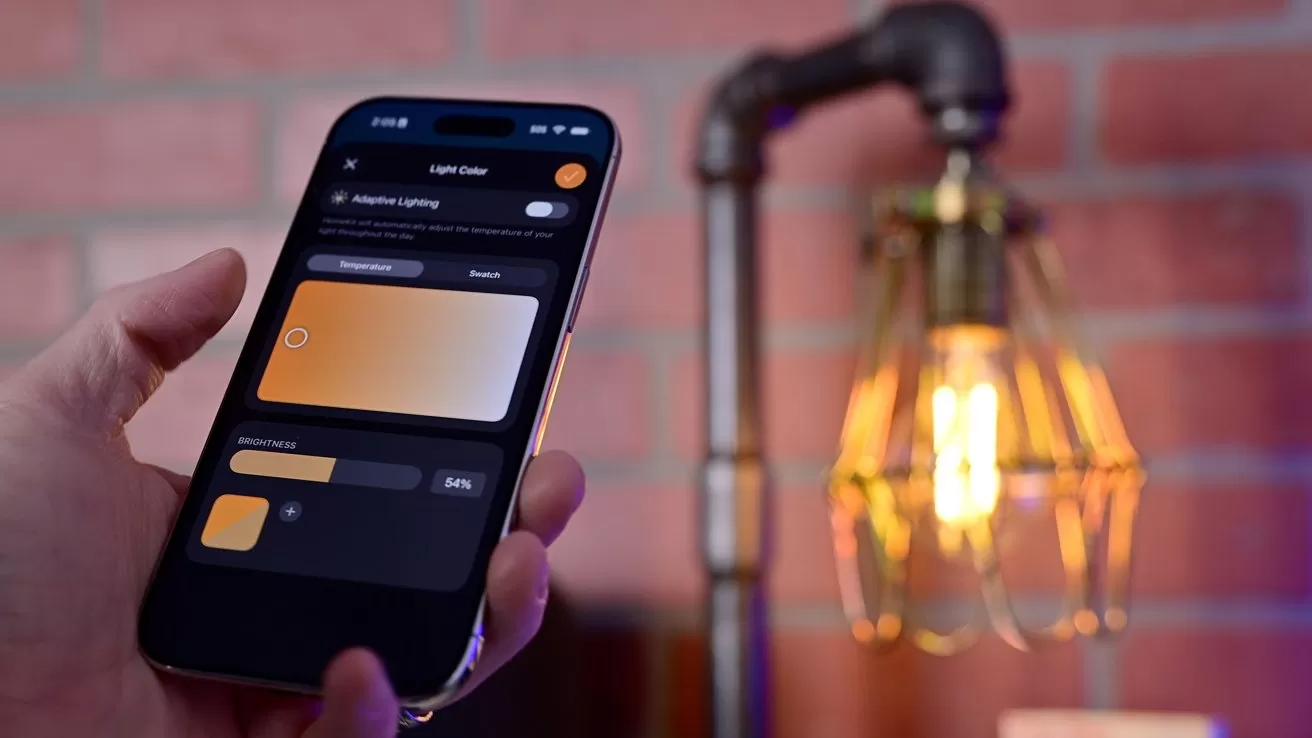 Hand holding a smartphone with a smart lighting app controlling warm light color and brightness, next to an exposed vintage-style bulb lamp against a red brick wall