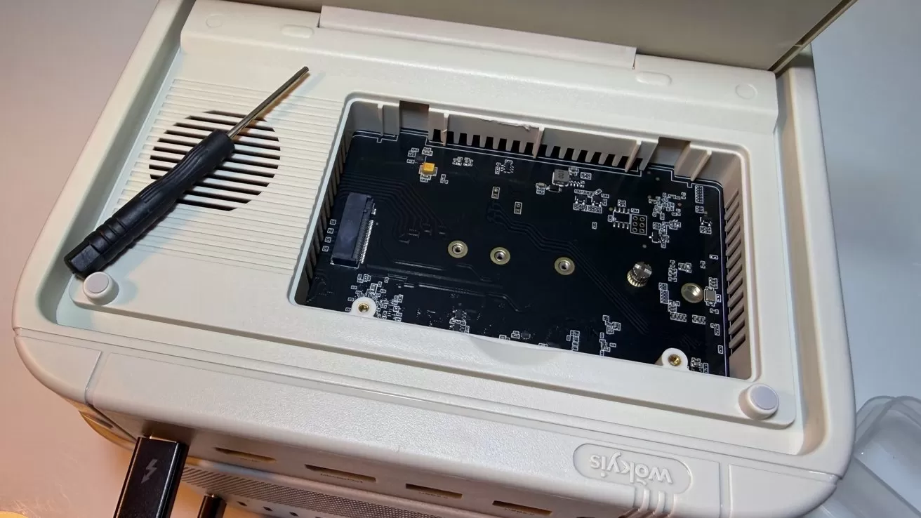 Open beige electronic device with top cover lifted, exposing a black circuit board inside and a small screwdriver resting beside the compartment near a speaker-style grille