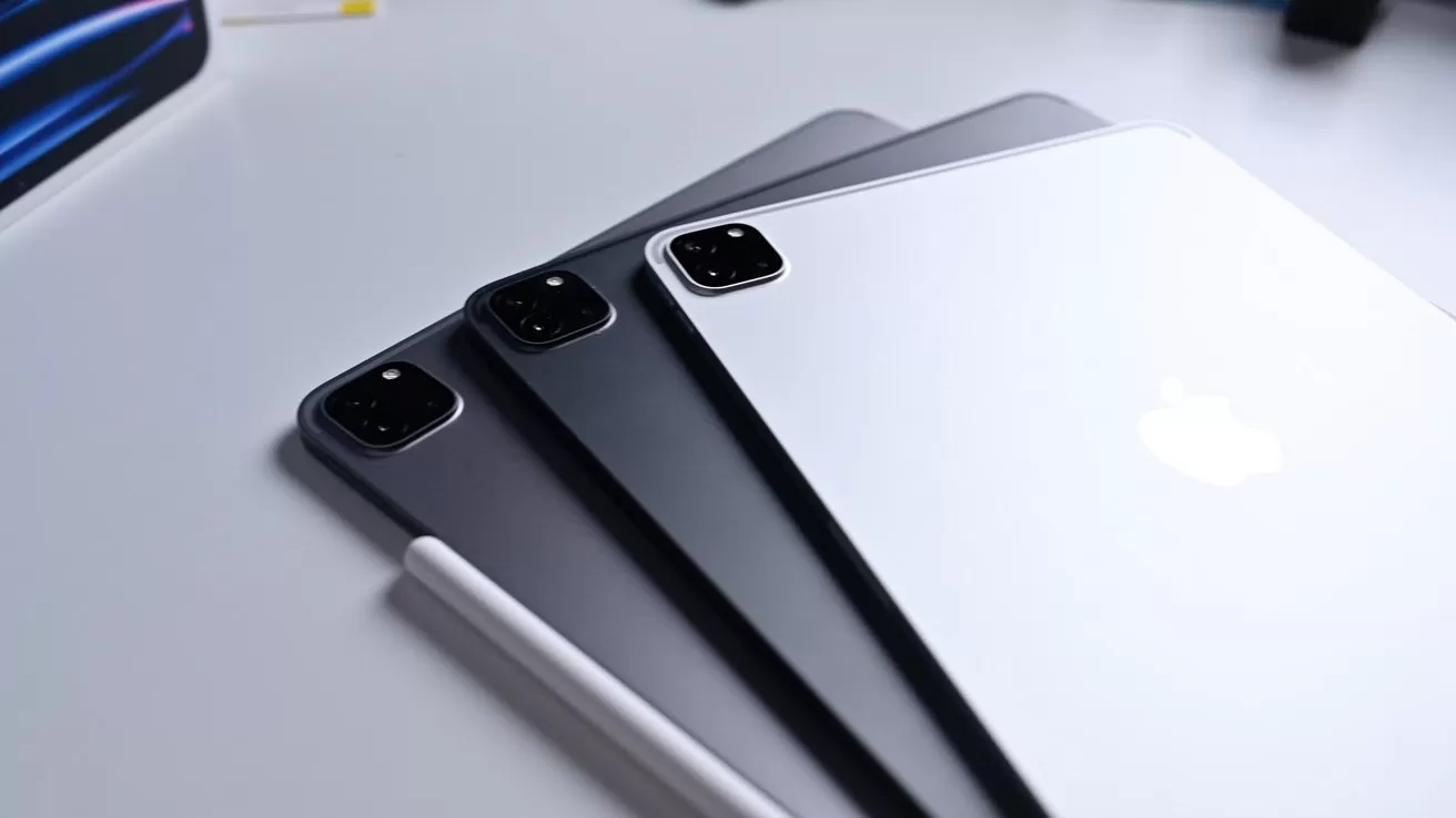 Three Apple iPad tablets in different colors stacked diagonally on a white surface, showing rear dual cameras, with an Apple Pencil resting alongside the bottom tablet