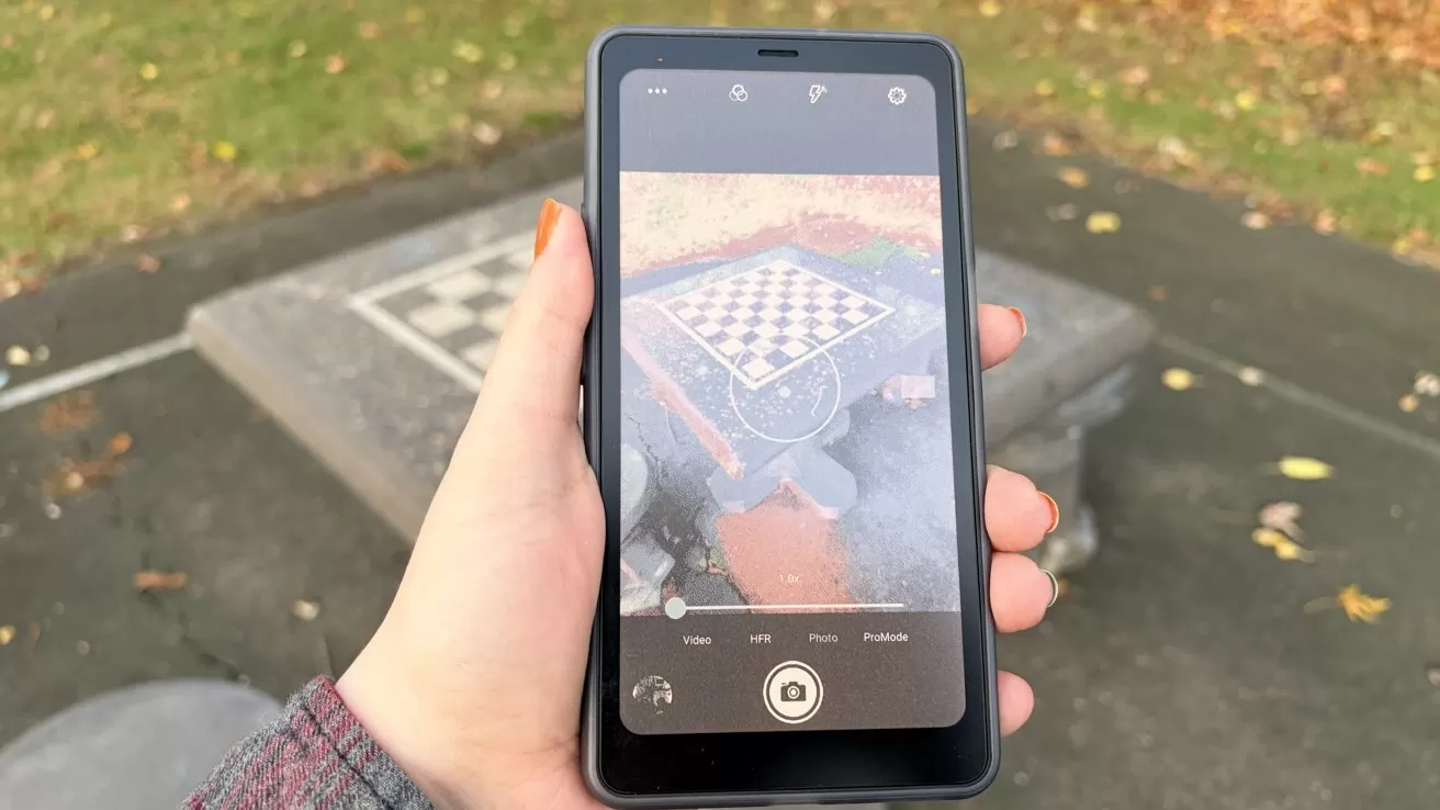 Hand holding a smartphone displaying a chessboard on a park table, with grass and fallen leaves visible in the background.