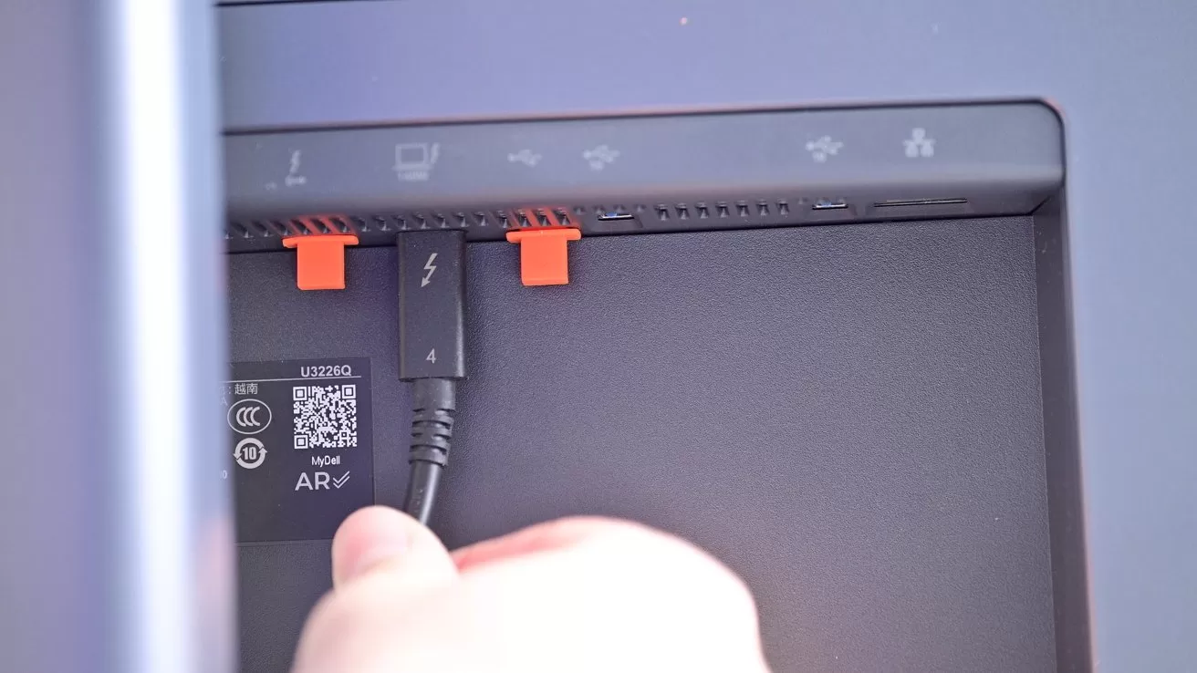 Hand plugging a Thunderbolt cable into a port on the back of a monitor or computer, near orange clips and a QR code label with small icons above the ports