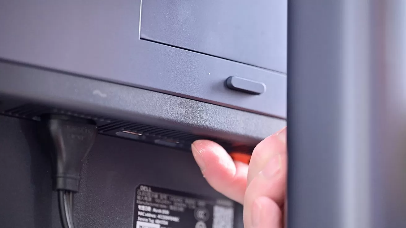 Hand reaching behind a monitor to press a small button near the power cable and label, suggesting adjusting settings or powering the device on or off