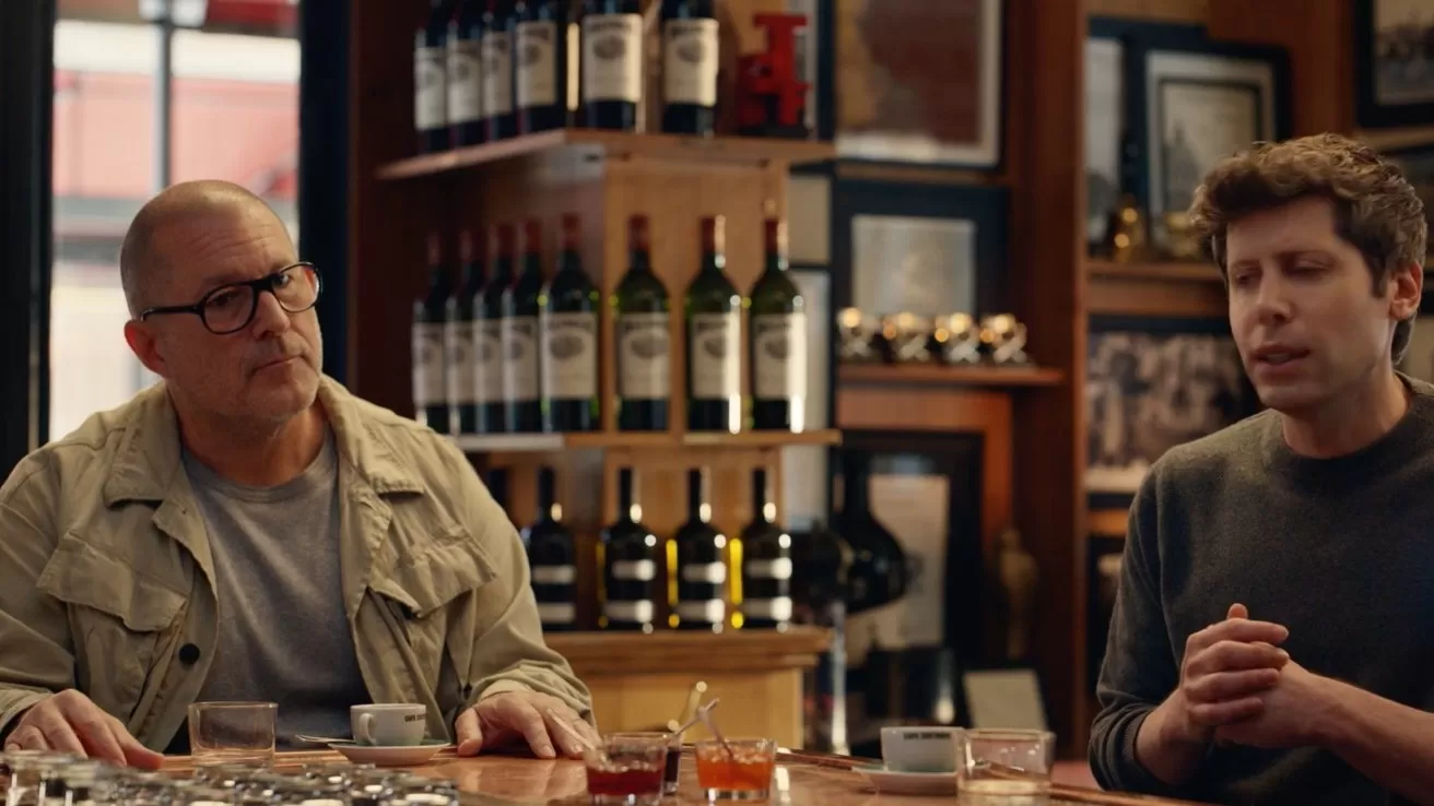 Two men sit at a bar counter with drinks, talking. Shelves of wine bottles and framed photos fill the background, creating a cozy, warmly lit, conversational setting.