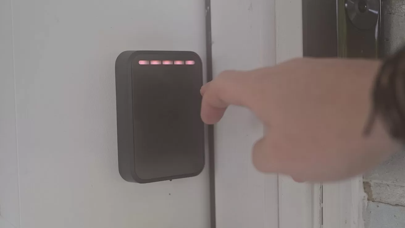 Hand reaching toward a wall-mounted electronic access panel with small red indicator lights, next to a closed door indoors