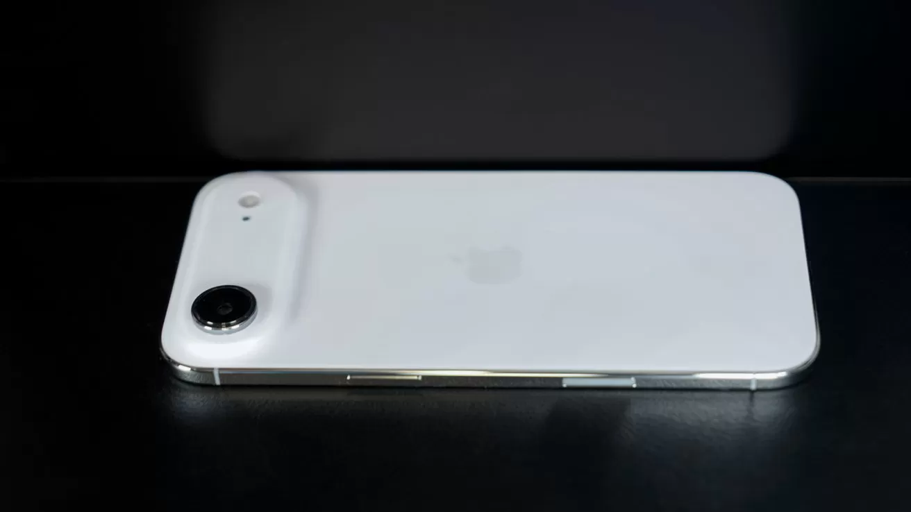 White smartphone lying face down on a dark surface, showing a single large rear camera lens and flash in the top left corner with a faint Apple logo in the center