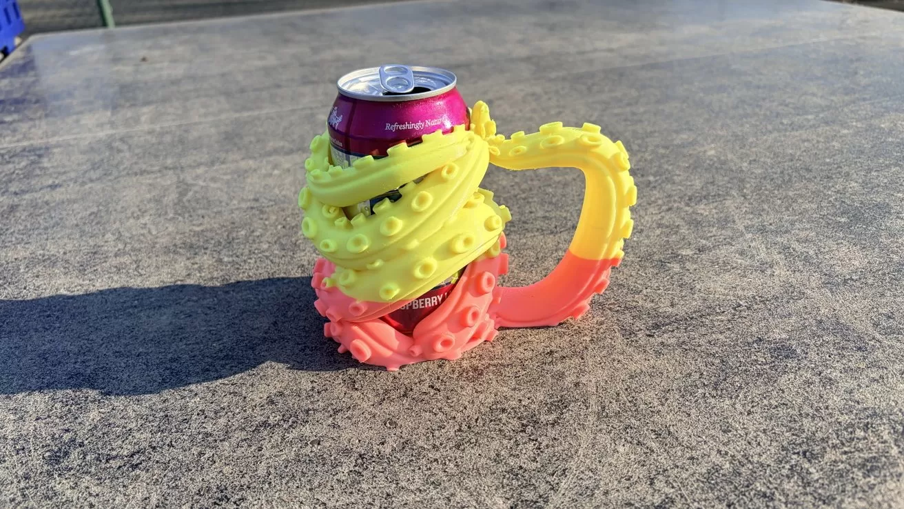 Colorful tentacle-shaped drink holder gripping a pink soda can on a concrete surface in bright sunlight, casting a long shadow across the ground