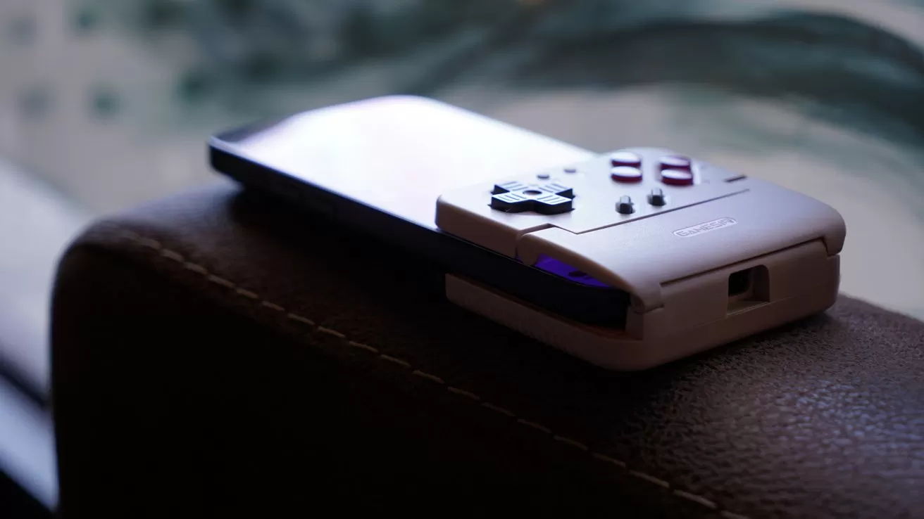 iPhone attached to a retro-style handheld gaming controller with D-pad and buttons, resting on a brown leather surface with soft, blurred background.