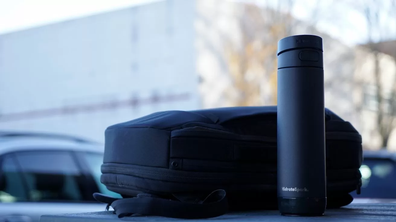 A black tumbler bottle and a dark backpack rest on a concrete surface with blurred outdoor background.