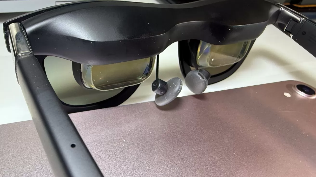 Black augmented reality glasses on a surface, with attached earbuds visible, alongside a metallic tablet or device in the background.
