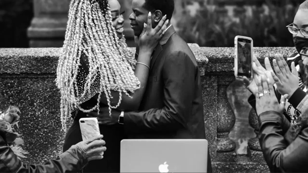 Couple embracing closely outdoors while friends take photos with phones; a laptop with an Apple logo sits in the foreground, all captured in black and white