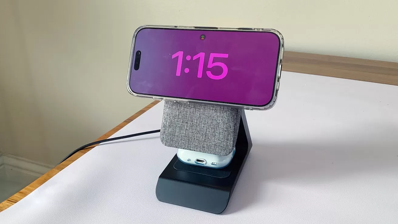 Smartphone in landscape mode displaying 1:15 with bright pink numbers, mounted on a gray fabric-covered charging stand holding wireless earbuds, on a white desk against a plain wall