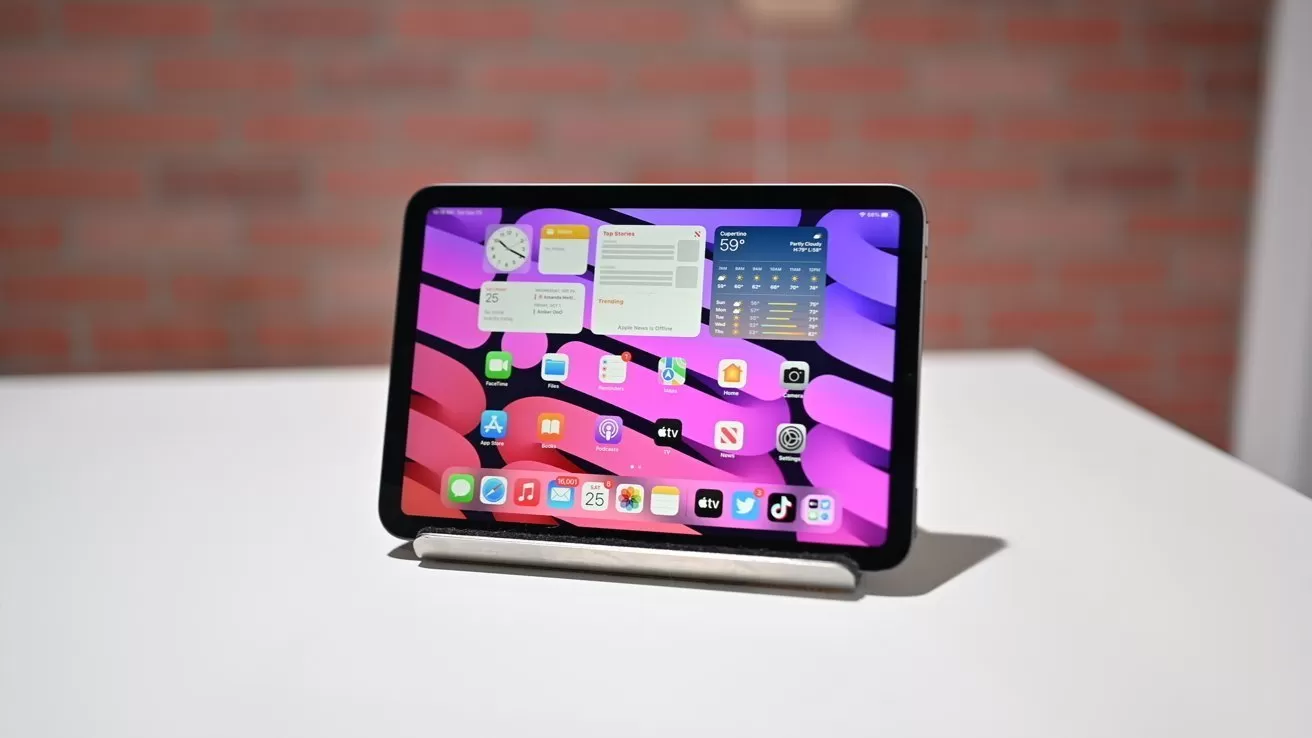 Tablet on a stand displaying a colorful pink and purple home screen with app icons and widgets, placed on a white table in front of a blurred brick wall background