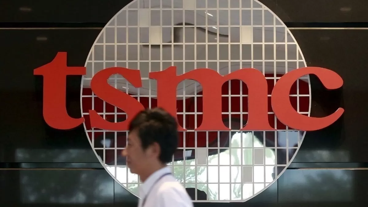 Person walking past large red TSMC logo on a dark wall, with a circular grid pattern behind the letters suggesting a semiconductor wafer backdrop