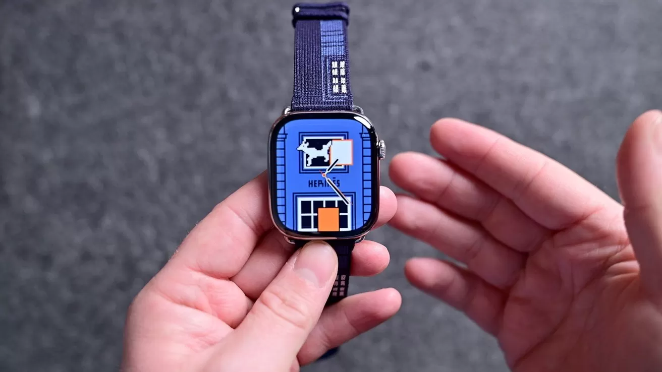 Hands holding a smartwatch with a blue band, displaying a bright blue watch face featuring a house-like graphic and bold shapes, against a gray textured background