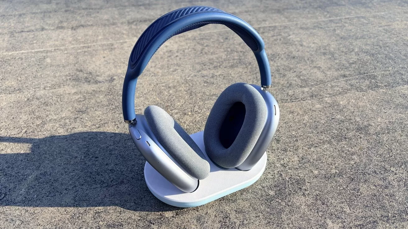 Blue over-ear wireless headphones with cushioned ear cups rest on a white oval charging stand, placed outdoors on a textured gray concrete surface in bright sunlight