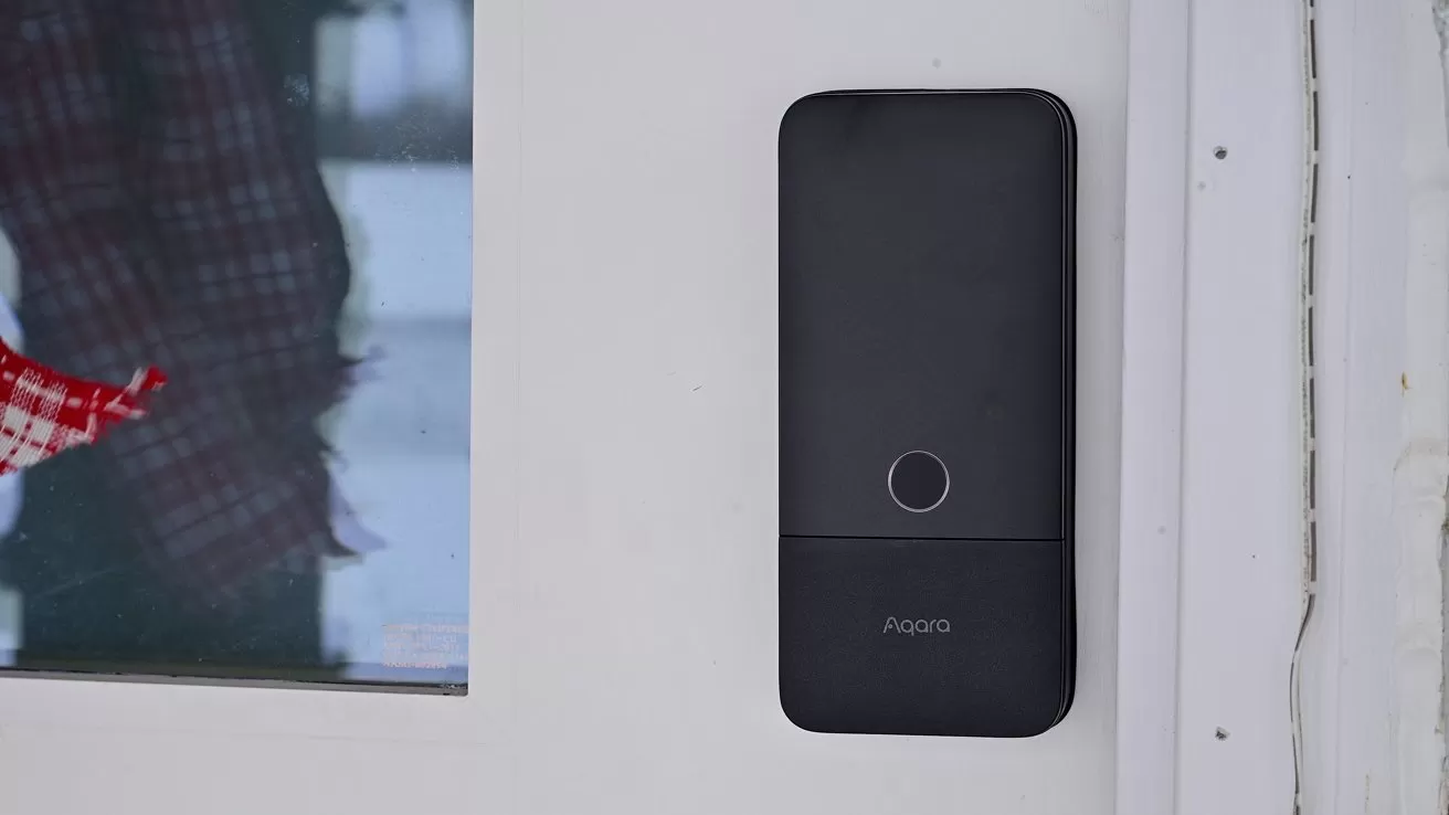Black rectangular smart doorbell with a circular button in the center mounted on a white wall beside a window showing a partial view of a person in plaid clothing