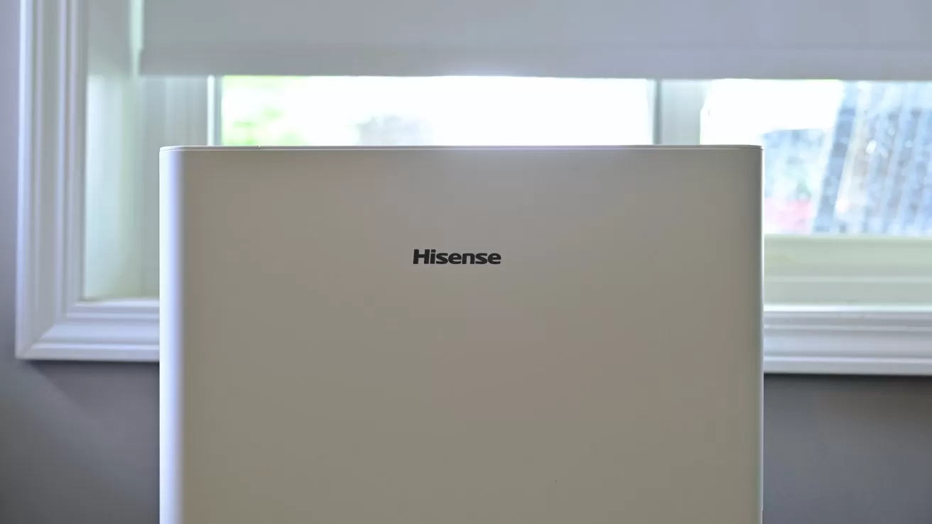 White Hisense appliance standing in front of a bright window with a white blind partially lowered, framed by simple white trim in a clean, modern room