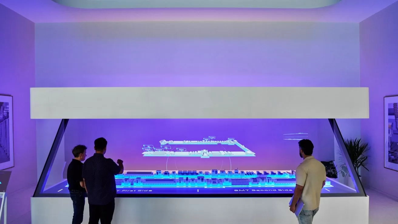 Three people stand around a large, futuristic interactive display table with glowing blue and purple lighting, viewing a digital architectural model in a modern, minimalistic room