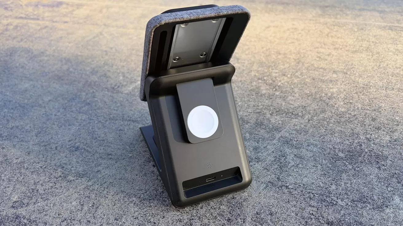 Black folding charging stand on rough gray surface, with fabric-covered phone rest, circular white charging pad for a smartwatch, and small USB-C port near the base