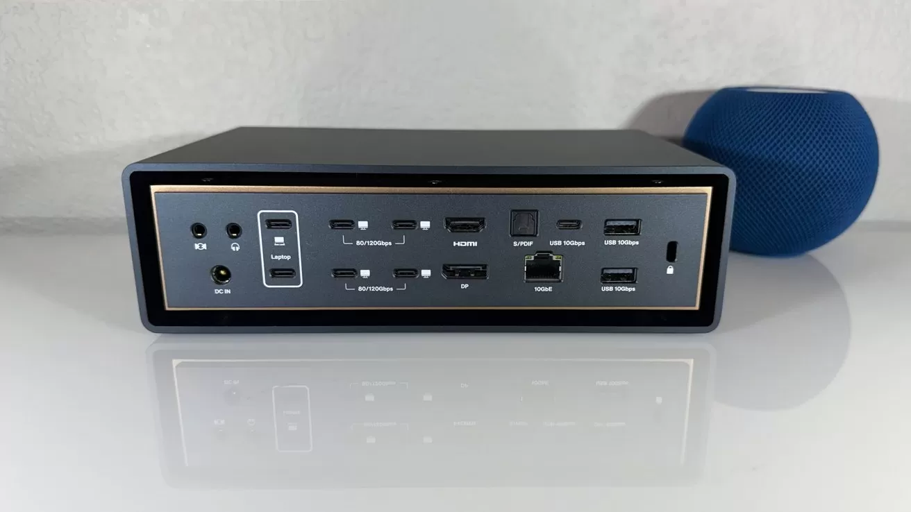 Rectangular docking station rear panel showing multiple USB-C, USB-A, HDMI, DisplayPort, audio jacks, Ethernet, power input, and S/PDIF ports, sitting on a white surface with a blue speaker behind