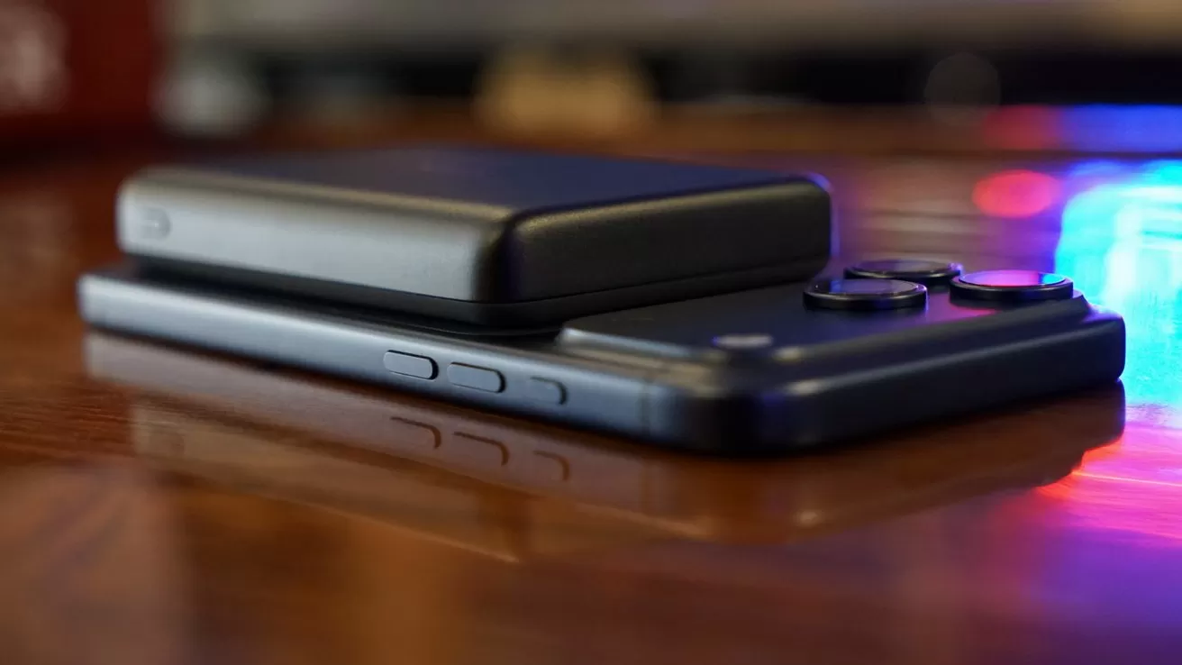 Portable power bank resting on an iPhone17 Pro Max, both lying on a polished wooden surface with soft colorful reflections in the background