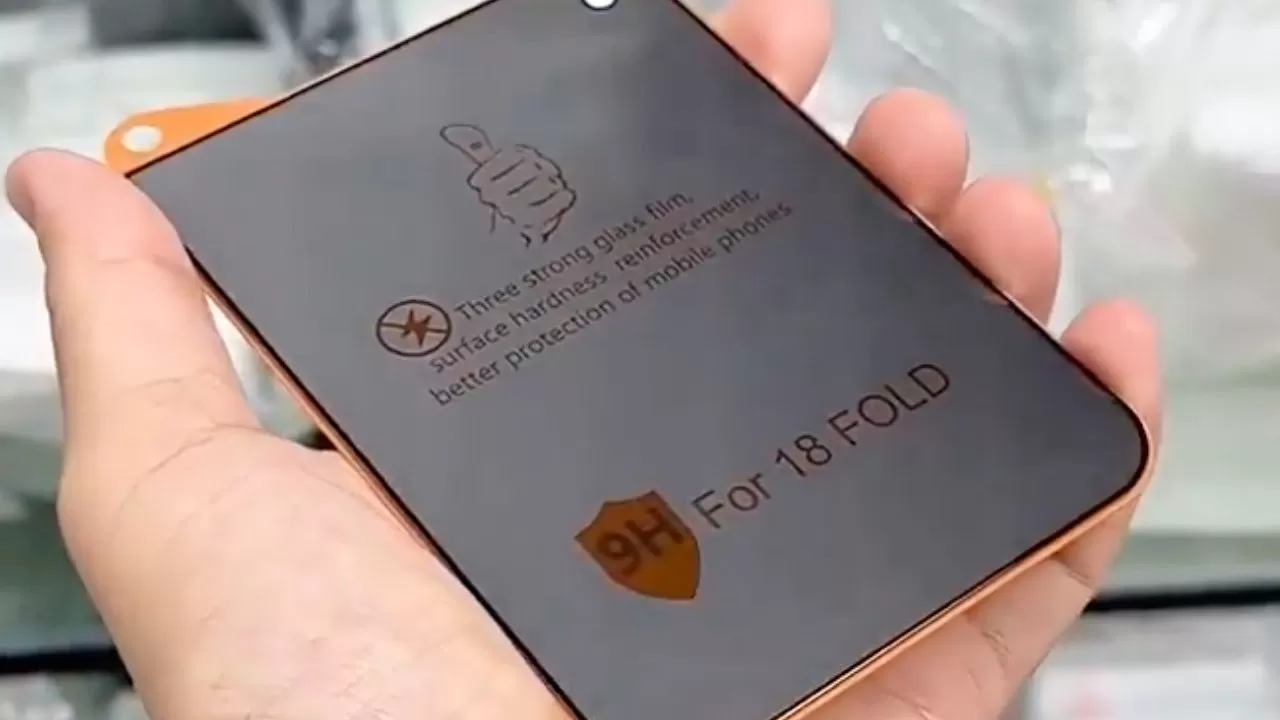 Hand holding a dark rectangular 9H screen protector labeled For 18 FOLD, with icons and small protective text, against a blurred indoor background.