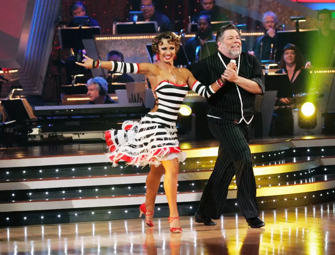 Smiling dance partners perform on a bright stage, woman in a striped ruffled dress and heels, man in dark suit, holding hands mid-step with band playing in background