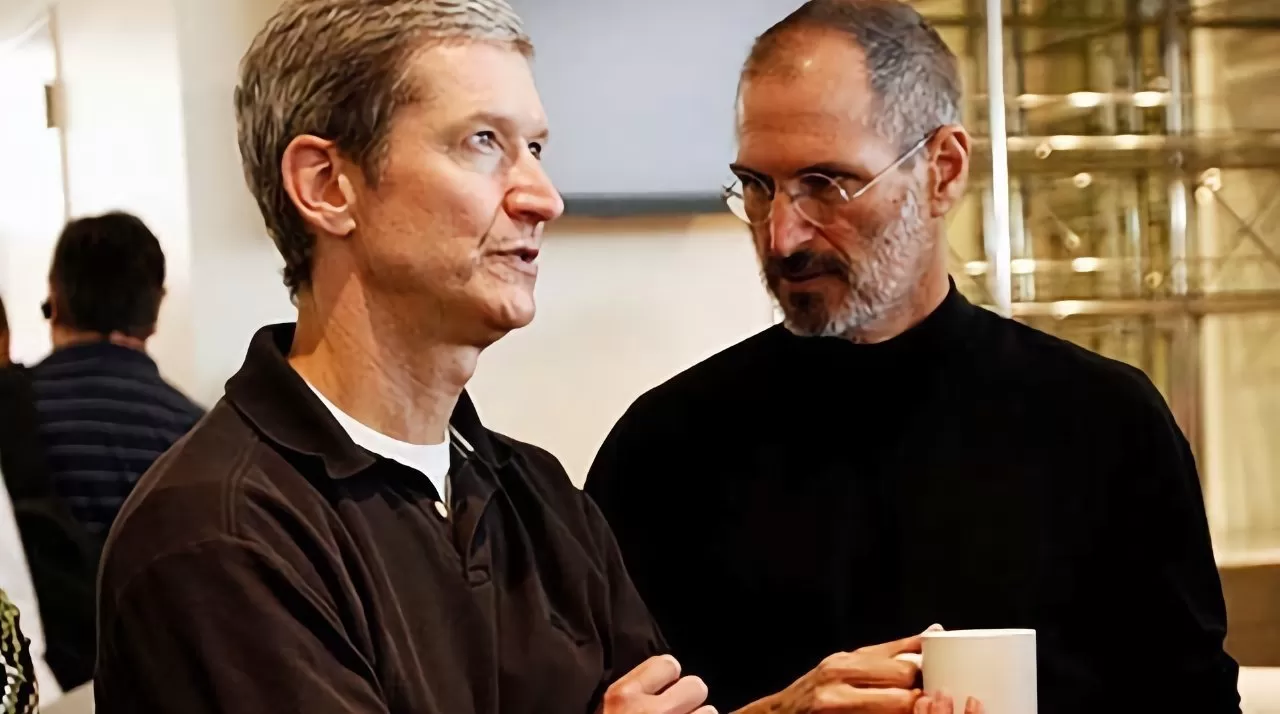 Two middleaged men stand close together indoors, conversing; one faces forward with arms crossed, the other in glasses and dark clothing holds a white mug, background slightly blurred