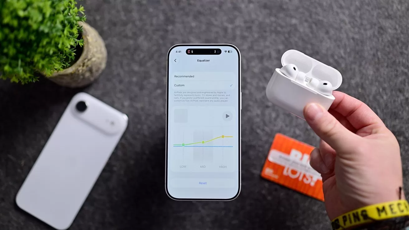 Hand holding open white wireless earbuds case beside a smartphone showing app stats on a gray surface, with another phone, small plant, and orange card in the background